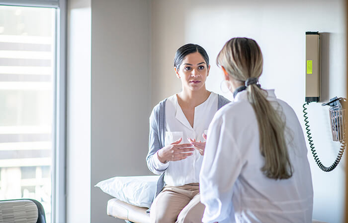 A young female of Indian ethnicity sits on the examination table at her gynaecologist's office. She is talking to her doctor about her health history.