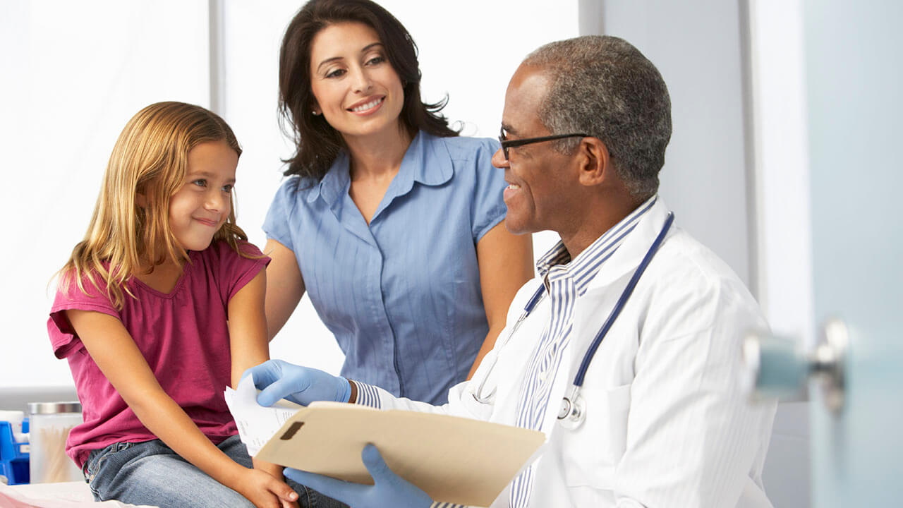 Young girl on exam table with mom standing behind her and male doctor is holding a patient file while looking at the girl