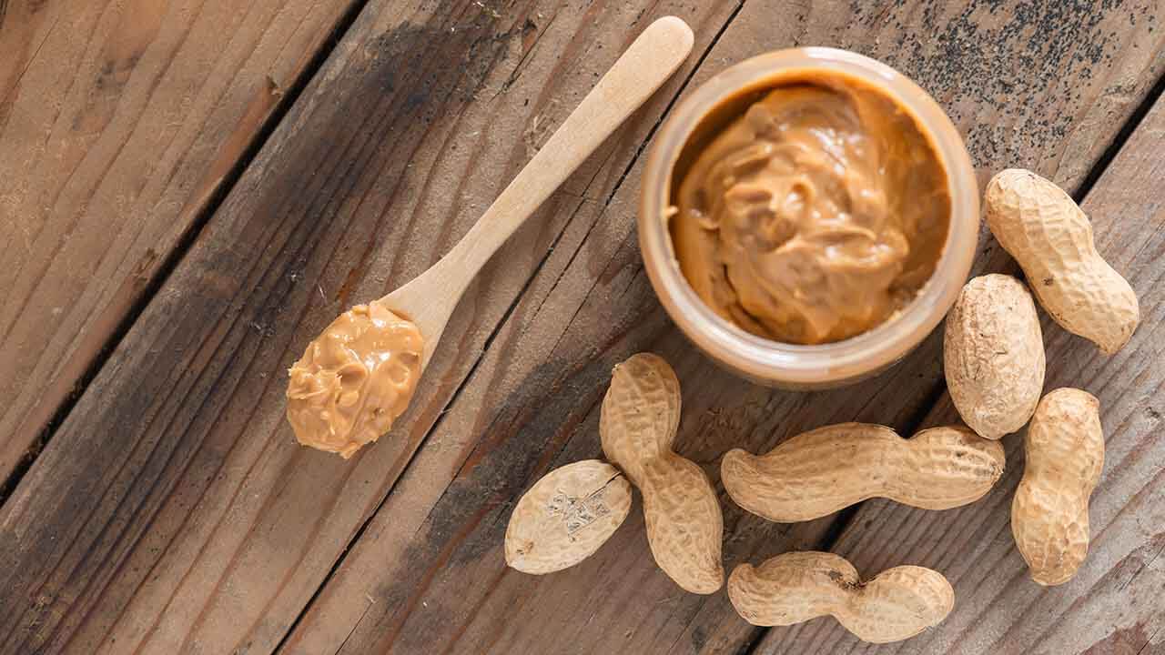 Peanut butter on a wooden spoon and in a jar on a wooden table next to peanuts in shells
