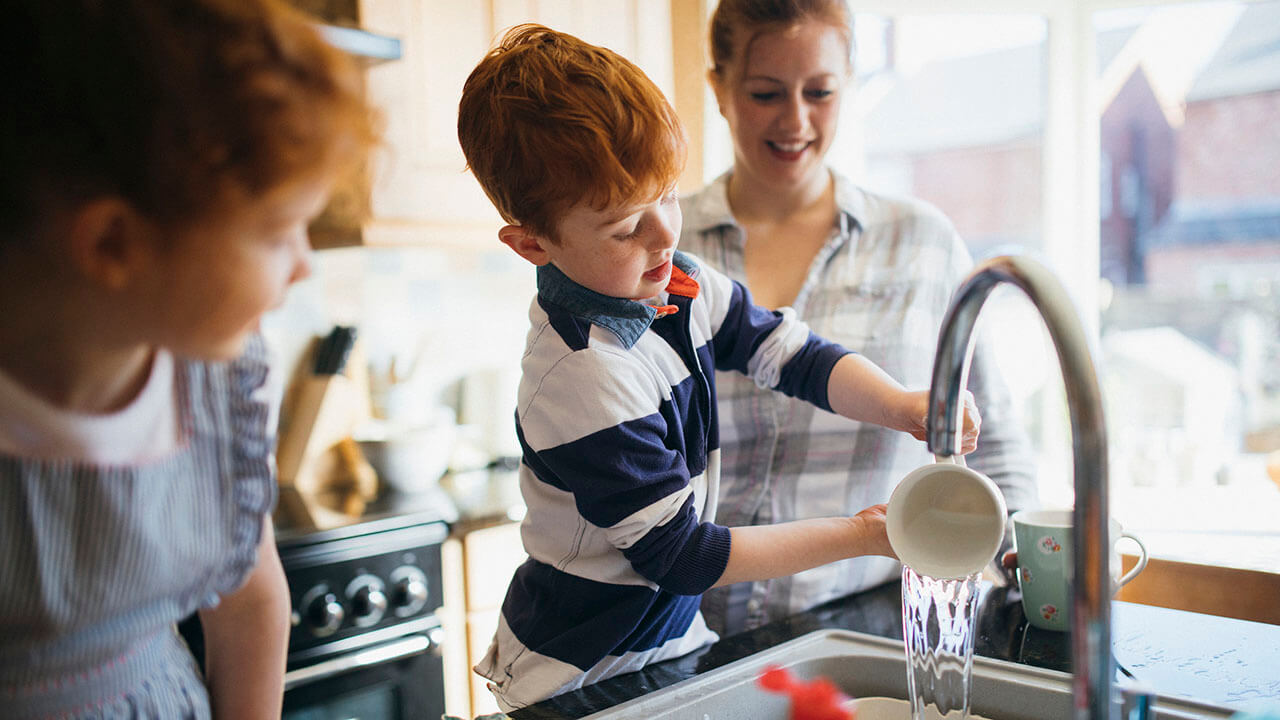 Two children washing dishes while mom is watching with a smile on her face