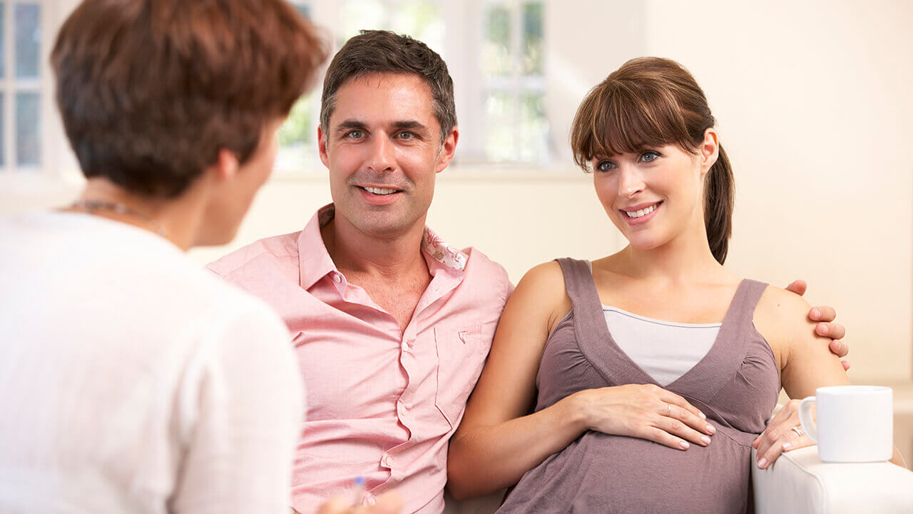 man and pregnant woman sitting on couch looking at a person