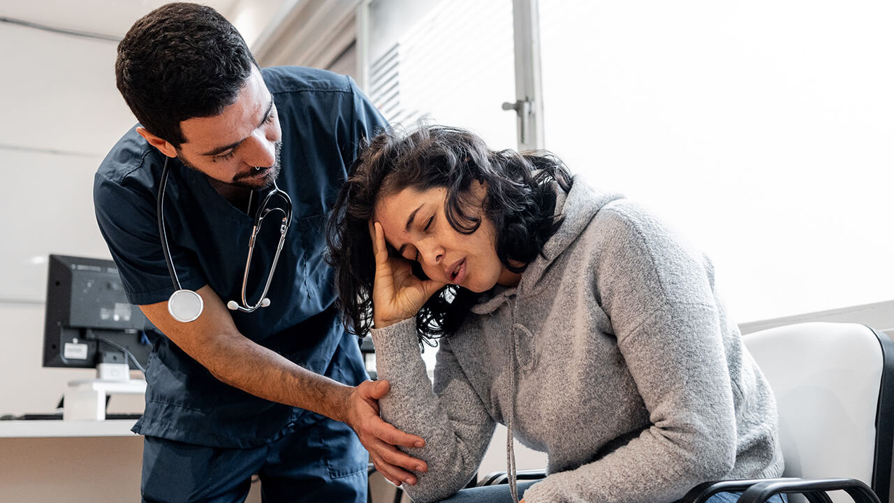 Young woman sitting in chair leaning forward with her hand on her head and a male nurse leans over her.