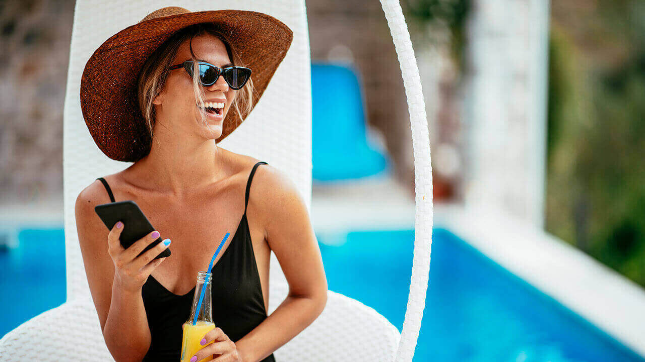 Woman in her bathing suit, hat and sunglasses laughing, holding her phone and beverage