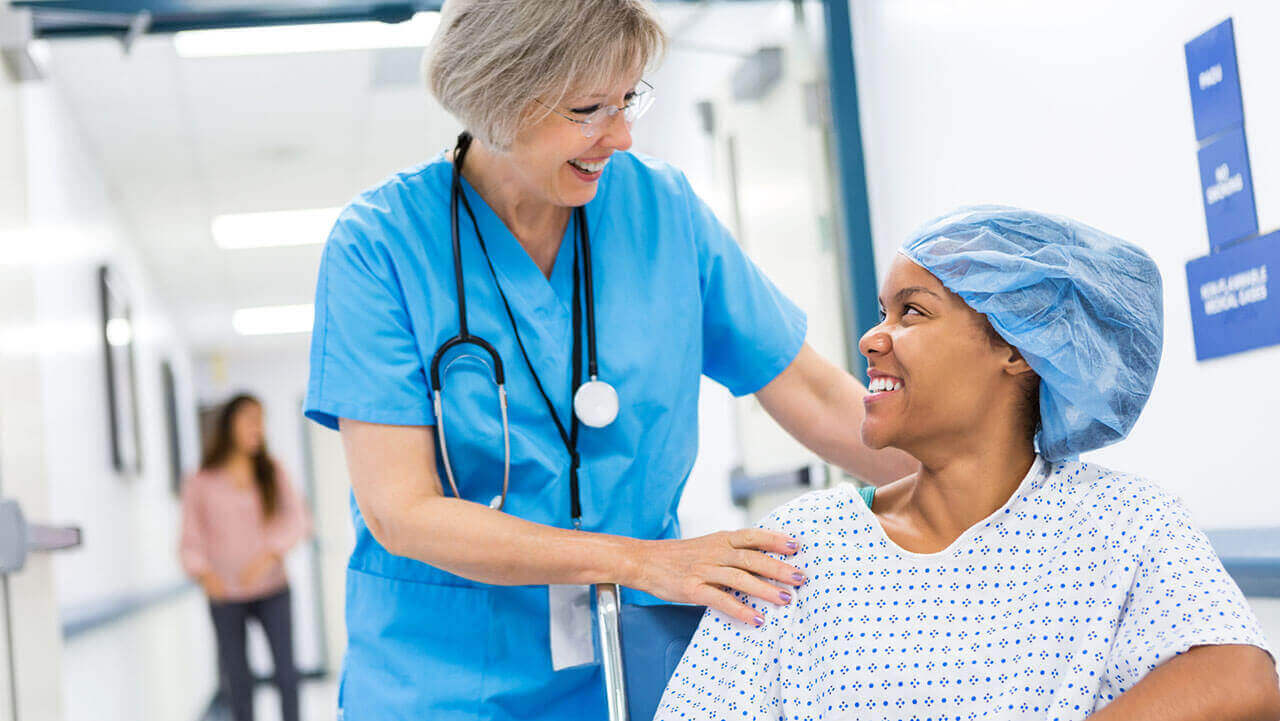 Female nurse pushing black woman in wheelchair prepped for surgery