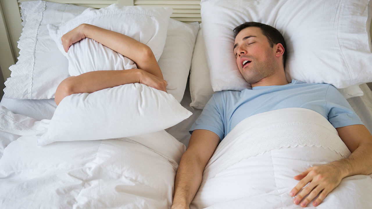 Woman covering her head with a pillow laying next to a male snoring in bed.