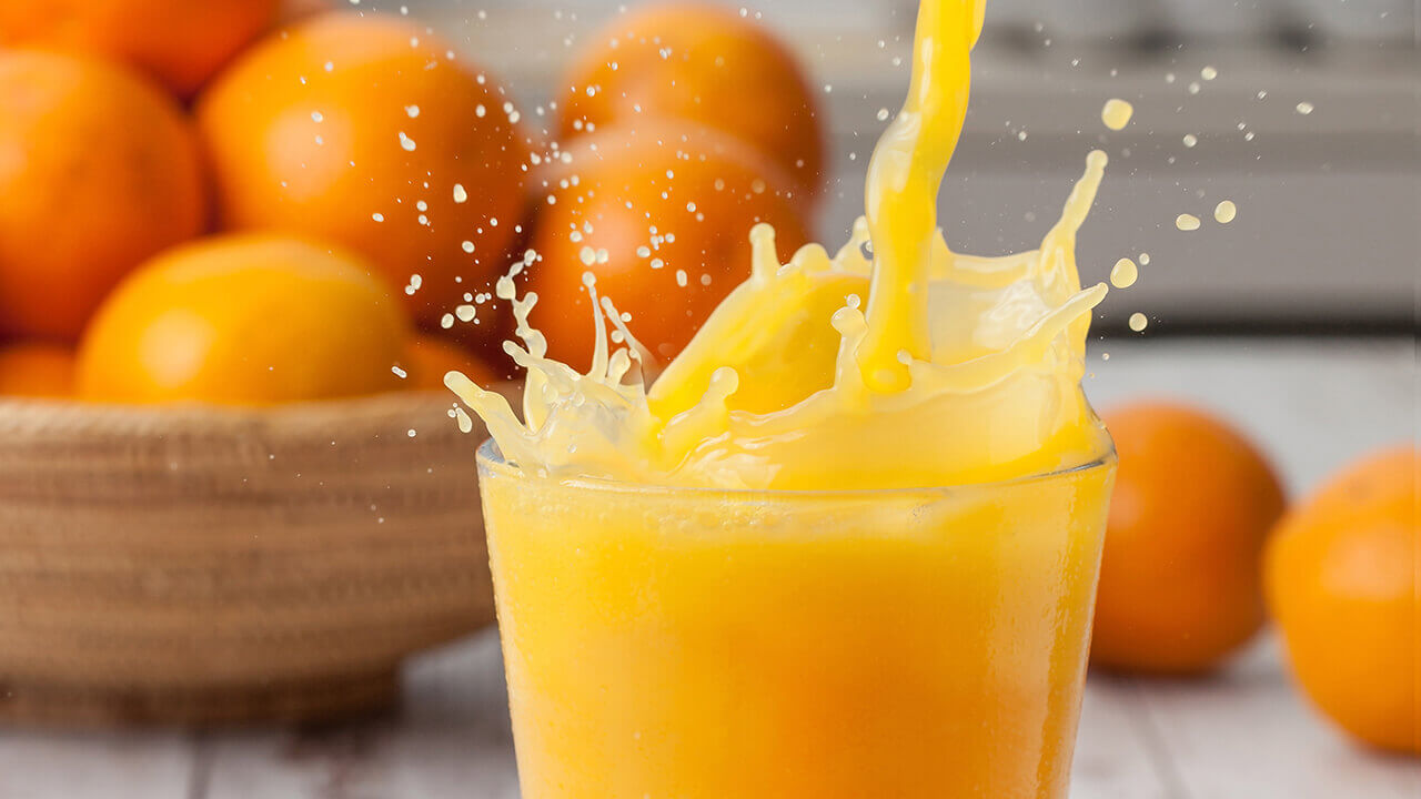 A glass of orange juice splashing out with a bowl of oranges in the background