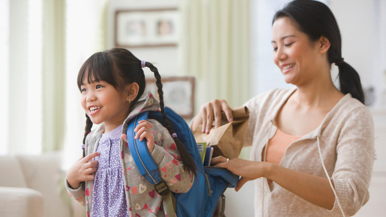 Young mom placing a lunch box in daughter's backpack