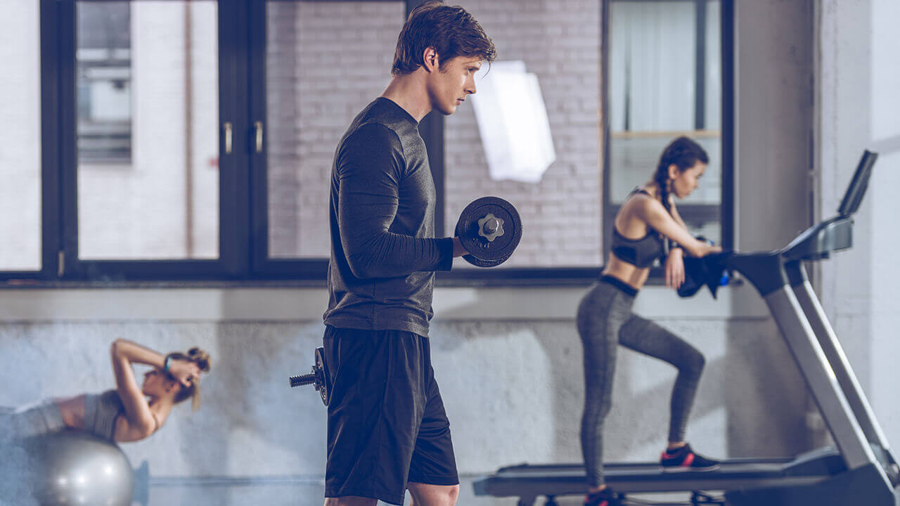 Man intensely lifting weights with a woman walking on a treadmill behind him