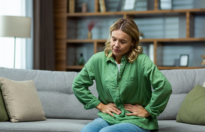 woman with blonde hair sits on gray couch with hands on midsection in pain